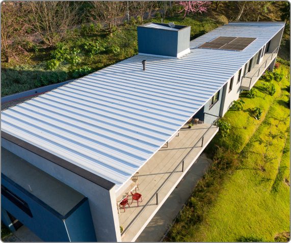 Vista aérea de uma residência contemporânea de telhado baixo coberto com isotelha trapezoidal clara, com placas fotovoltaicas em um trecho e vegetação ao redor.